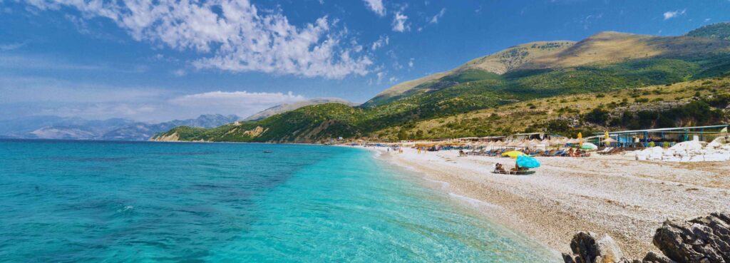 Albania budget travel guide featuring a scenic beach with turquoise water, sun loungers, and mountain views along the shore.