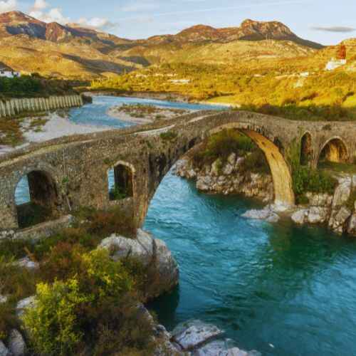 Ancient stone arch bridge crossing a turquoise river in the mountains, showcasing scenic landscapes for an Albania budget travel guide.